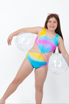 Front view of girl wearing the Cotton Candy Cut-Out rainbow tie-dye one-piece swimsuit with centre ring detail and side cut-outs while holding two beach balls.
