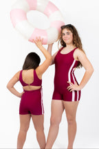 Front and back view of girls wearing The OG boy leg one-piece swimsuit in cherry maroon with pink side stripe detail, round scoop neckline, gold clasp back closure, and full coverage fit.