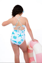 Back view of girl wearing a seashell and starfish one-piece swimsuit with adjustable tie-back straps and full coverage fit while leaning on a pink and white inflatable.