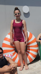 Front view of girl wearing The OG boy leg one-piece swimsuit in cherry maroon with pink side stripe detail while leaning on an orange and white inflatable ring by the pool.