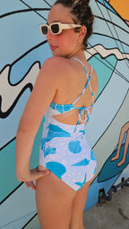 Back view of girl wearing a seashell and starfish one-piece swimsuit with adjustable tie-back straps and sunglasses.