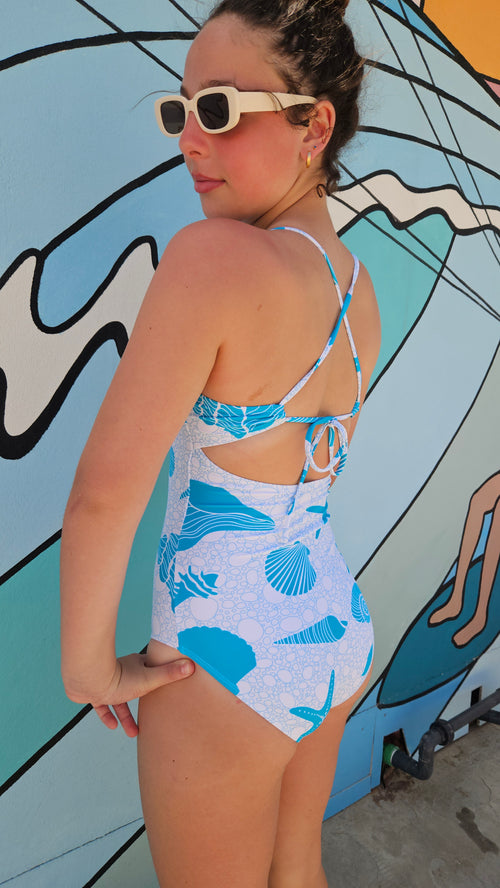 Back view of girl wearing a seashell and starfish one-piece swimsuit with adjustable tie-back straps and sunglasses.