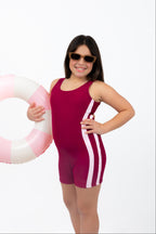 Front side view of girl wearing The OG boy leg one-piece swimsuit in cherry maroon with pink side stripe detail while holding a pink and white inflatable ring.