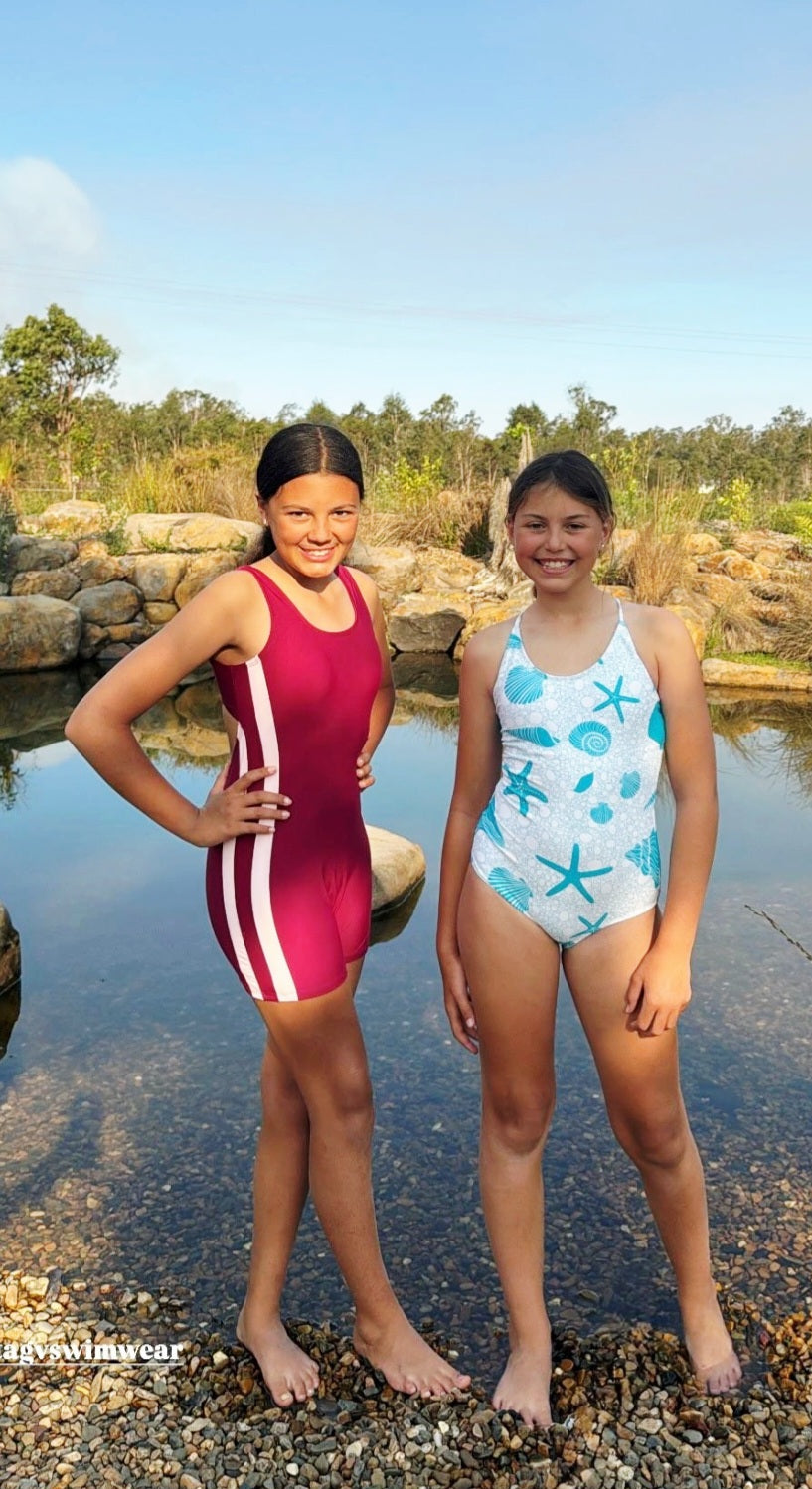 Girl wearing The OG boy leg one-piece swimsuit with pink side stripe detail standing beside girl wearing the Seafoam Sweetie swimsuit in front of a billabong.