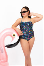 Front view of girl wearing the Coastal Bloom floral one-piece swimsuit with visible side ruching detail while leaning on a pink flamingo inflatable.
