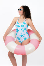 Girl wearing a seashell and starfish one-piece swimsuit with adjustable tie-back straps and full coverage fit while holding a pink and white inflatable